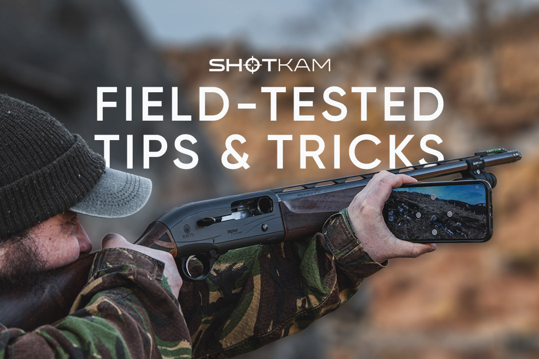 Field shooter using a shotgun equipped with a ShotKam camera, reviewing shot performance on a smartphone during a hunt.