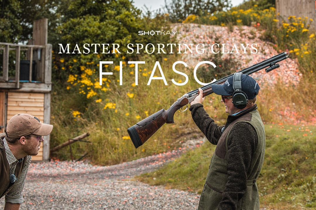Man in ear protection and shooting vest showing shotgun with ShotKam during FITASC practice on a clay-covered range.